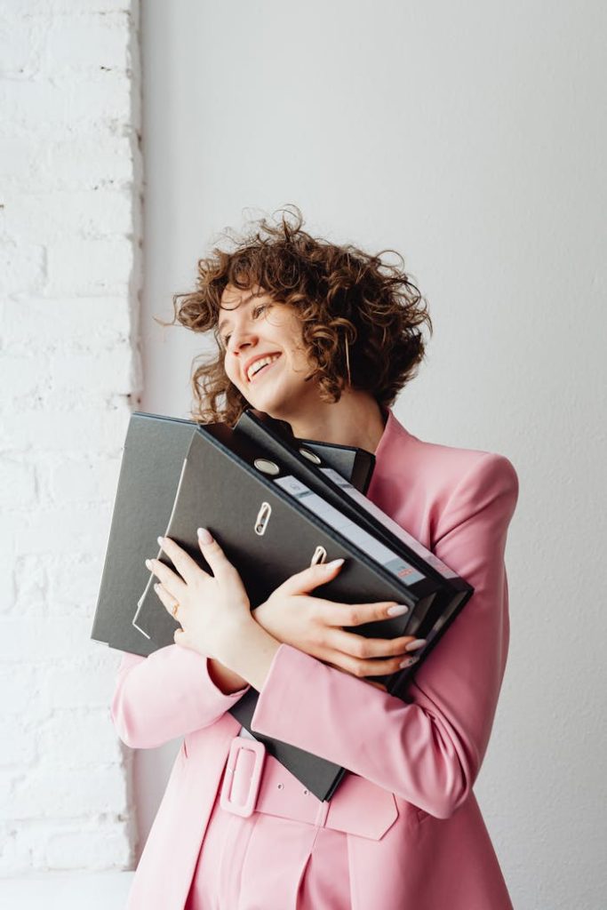 Cheerful woman embracing binders, wearing a pink blazer indoors with a bright smile.